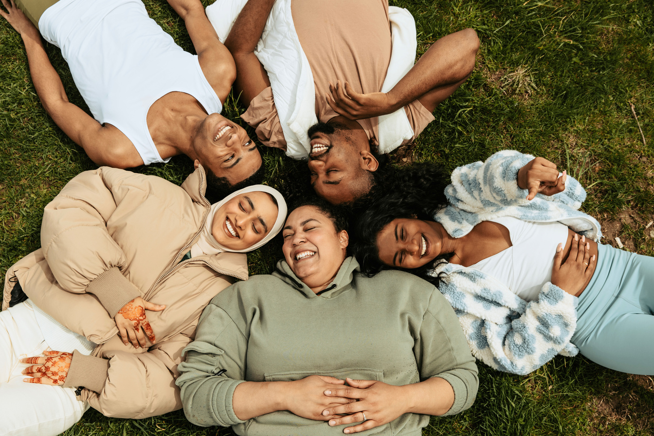 Multiracial and multi-faith group of young people laying on their backs in the grass, smiling and laughing with each other.